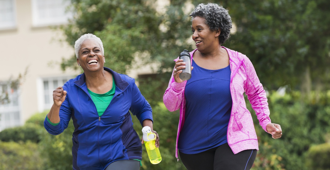Two senior African American women getting in shape together.  They are jogging or power walking on a sidewalk in a residential neighborhood, talking and laughing.